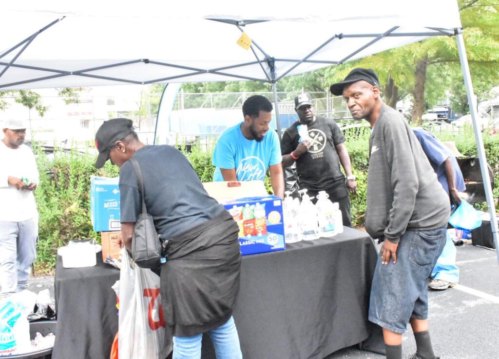 Volunteers distribute food and supplies at a community service event, with several individuals engaged and interacting under a tent.