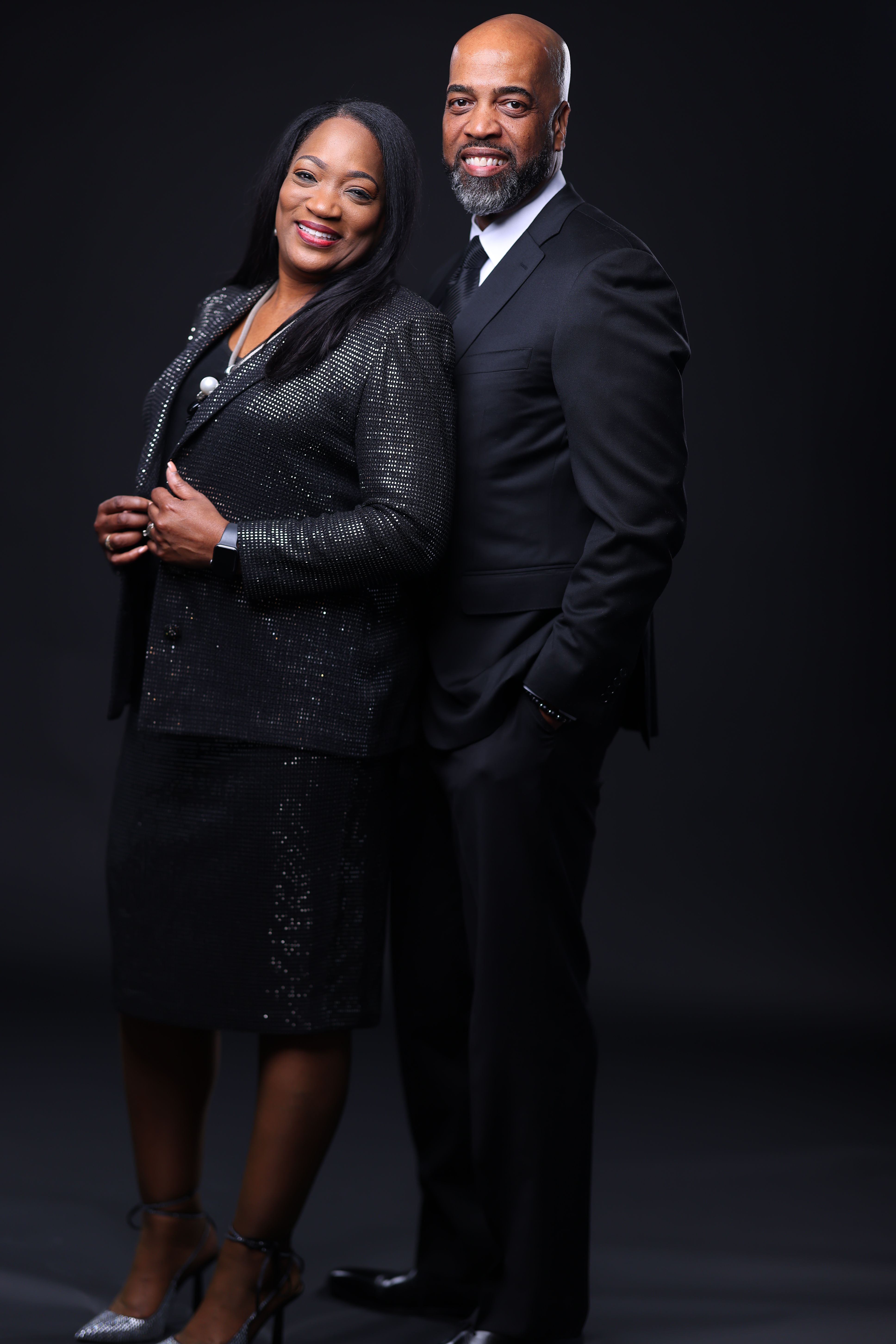 A confident couple poses together, both in formal black attire, exuding professionalism and elegance against a dark backdrop.