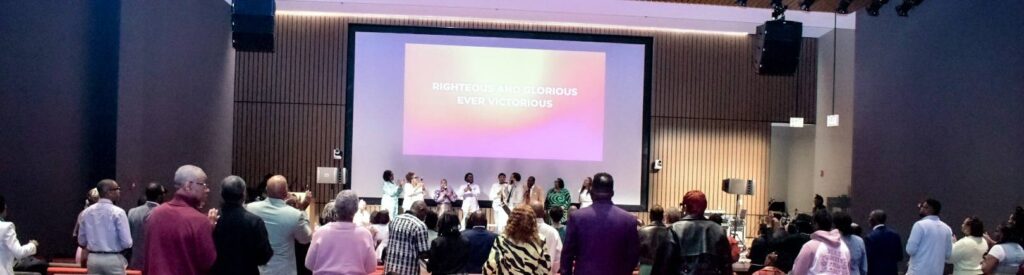 A congregation worships during a service, facing a screen displaying "Righteous and Glorious, Ever Victorious." The atmosphere is vibrant and uplifting.