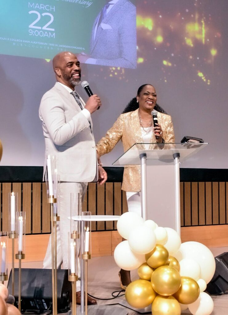 A man and woman stand at a podium, smiling and speaking to an audience during a celebration. Behind them, a backdrop displays an event date and details.